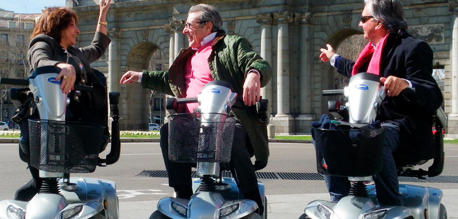 Tres personas en scooter de movilidad con la puerta de Alcalá de Madrid de fondo