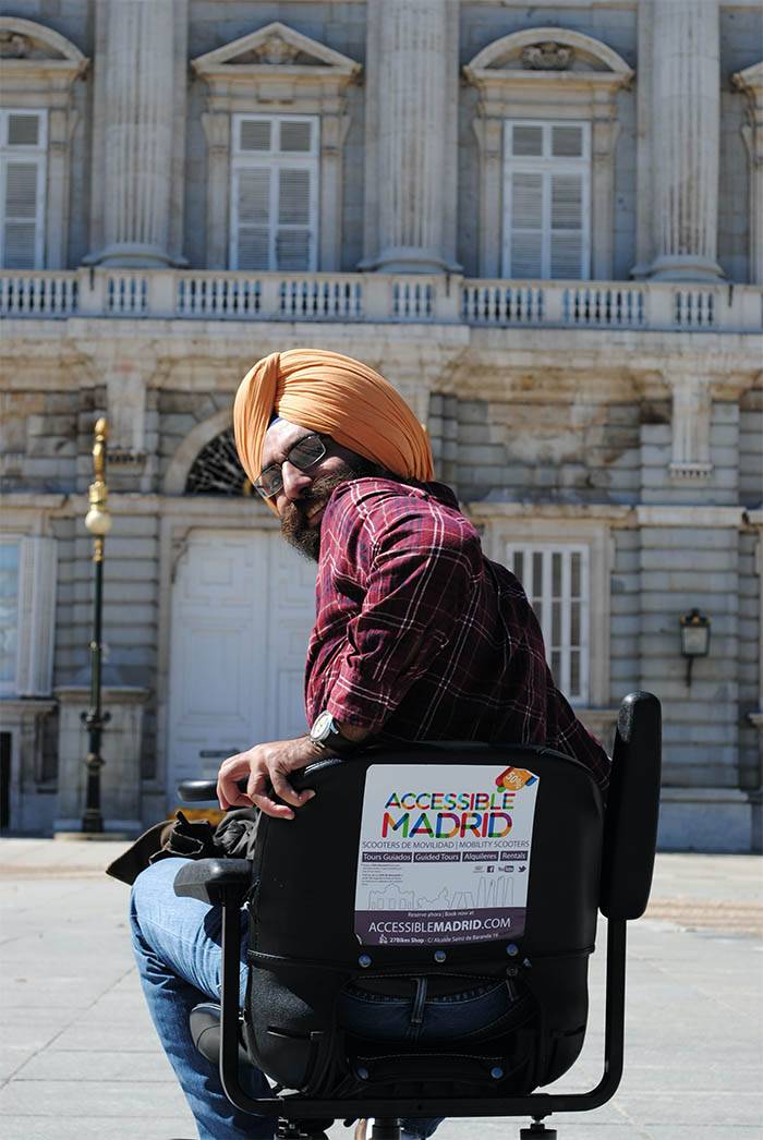 A person with a turban and Indian appearance poses sitting in a mobility scooter. In the background, the Royal Palace of Madrid